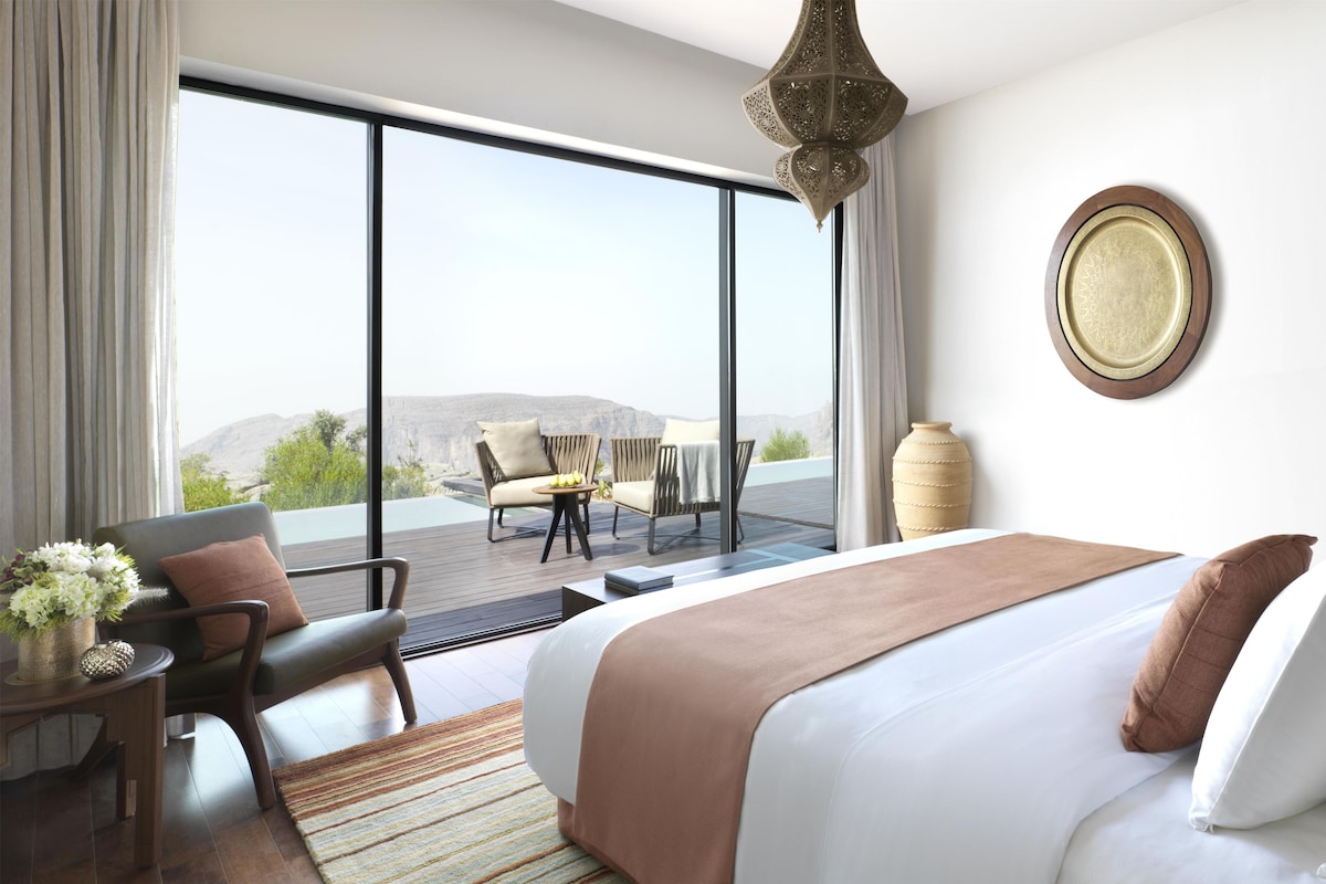 A hotel room with a large bed, a view of the outdoors, a dining area, and a decorative wall piece.