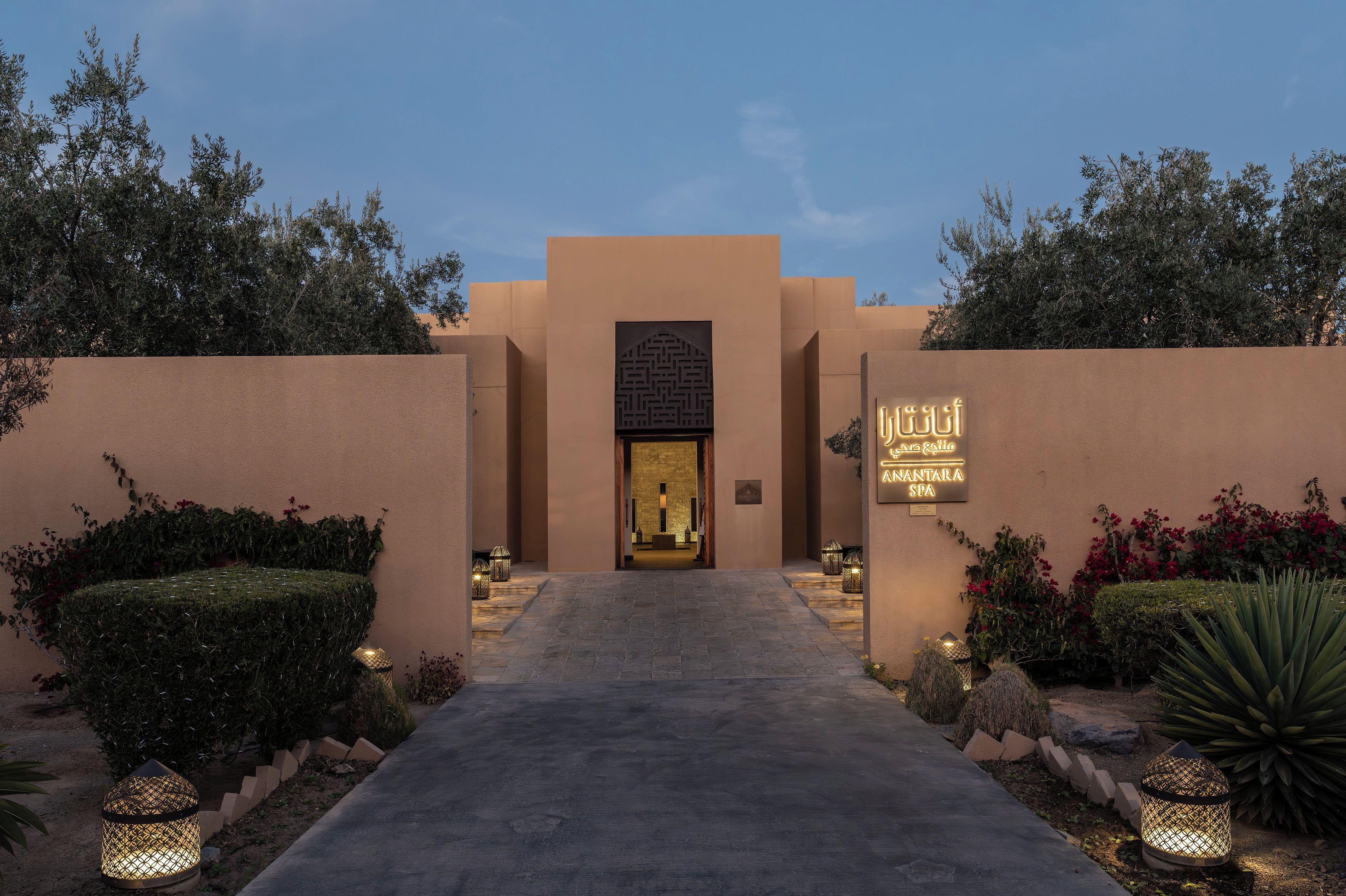 A hotel entrance with a pathway leading to the main building, surrounded by landscaped gardens and decorative lighting.