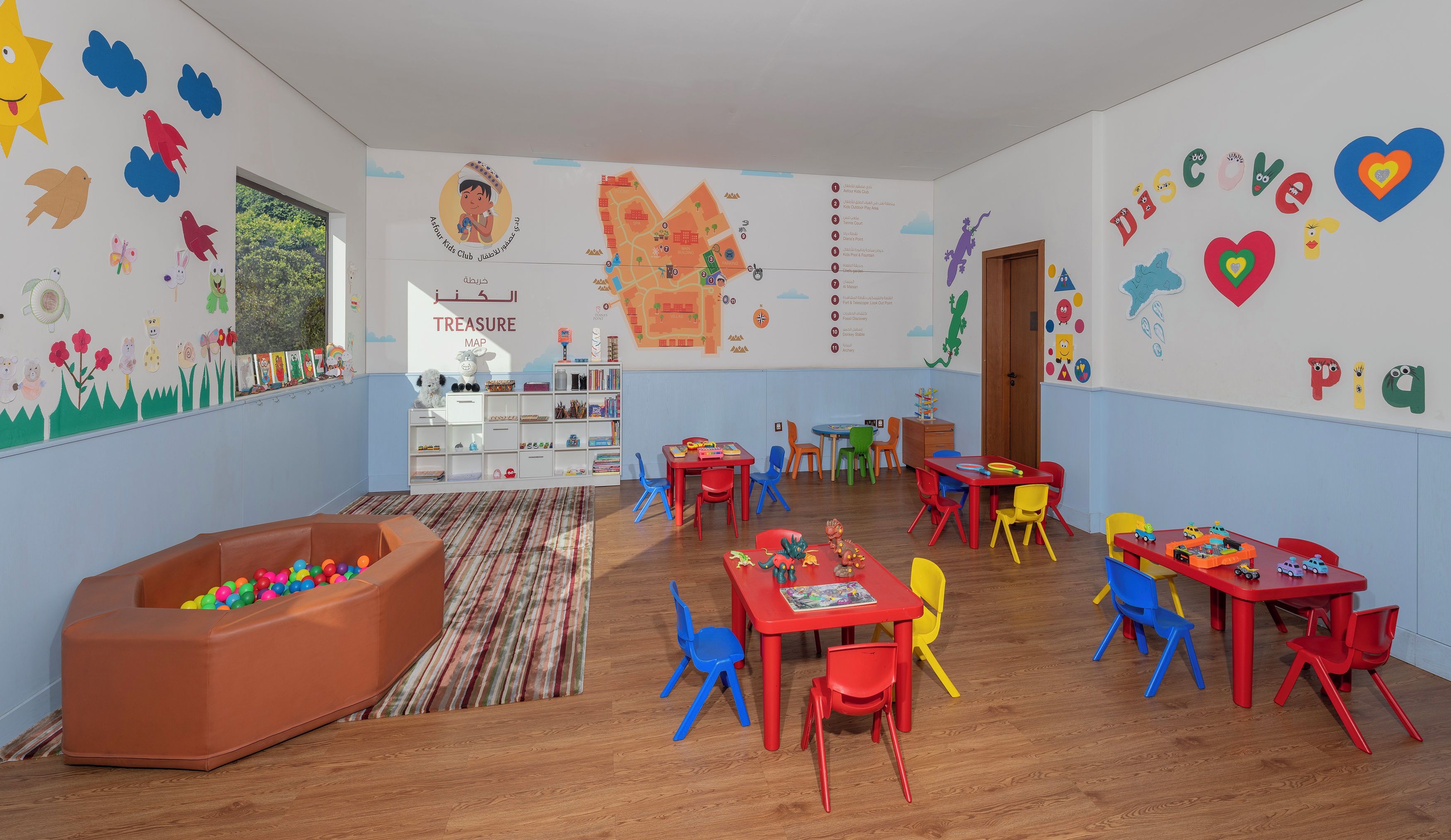 Una sala de juegos infantiles con mobiliario colorido, un mural con un mapa del tesoro y una piscina de pelotas.