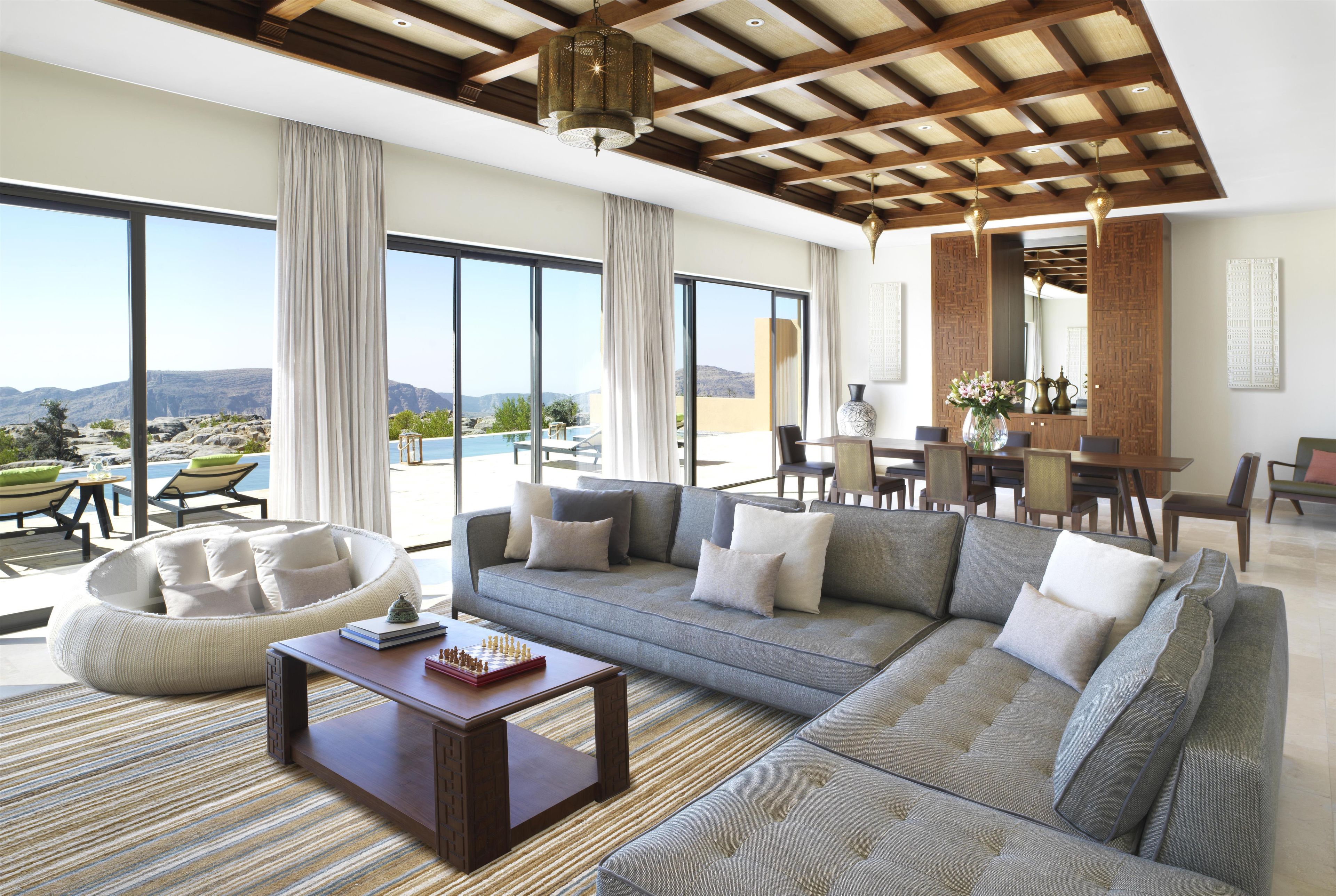 A spacious living area with a large sofa, a coffee table, and a dining table. There are large windows offering a view of the outdoors.