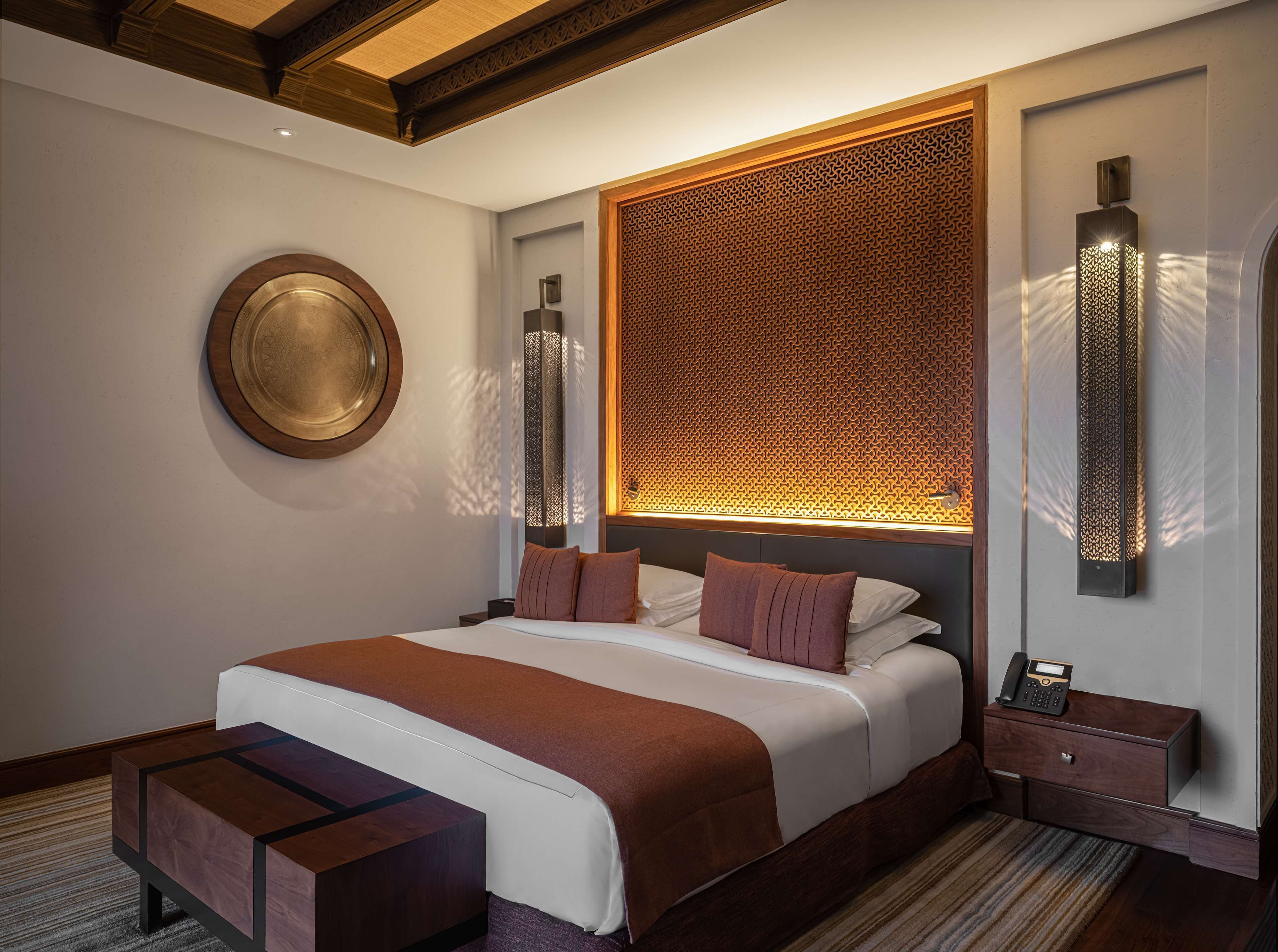 A modern hotel room with a large bed, a round mirror, a decorative wall panel, and a bedside table with a phone.
