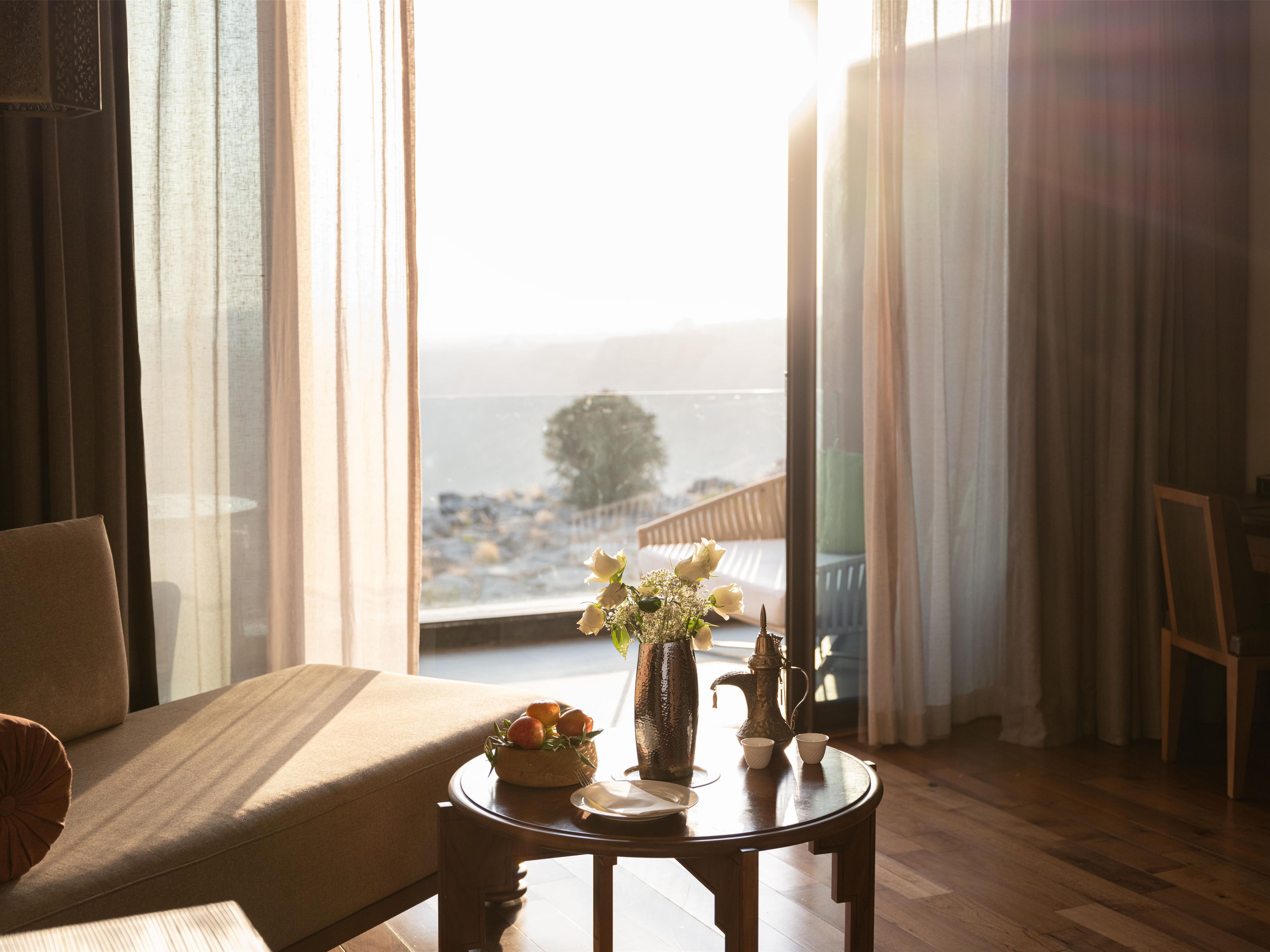 A room with a bed, a table with a vase of flowers, and a view of the sea through a window.