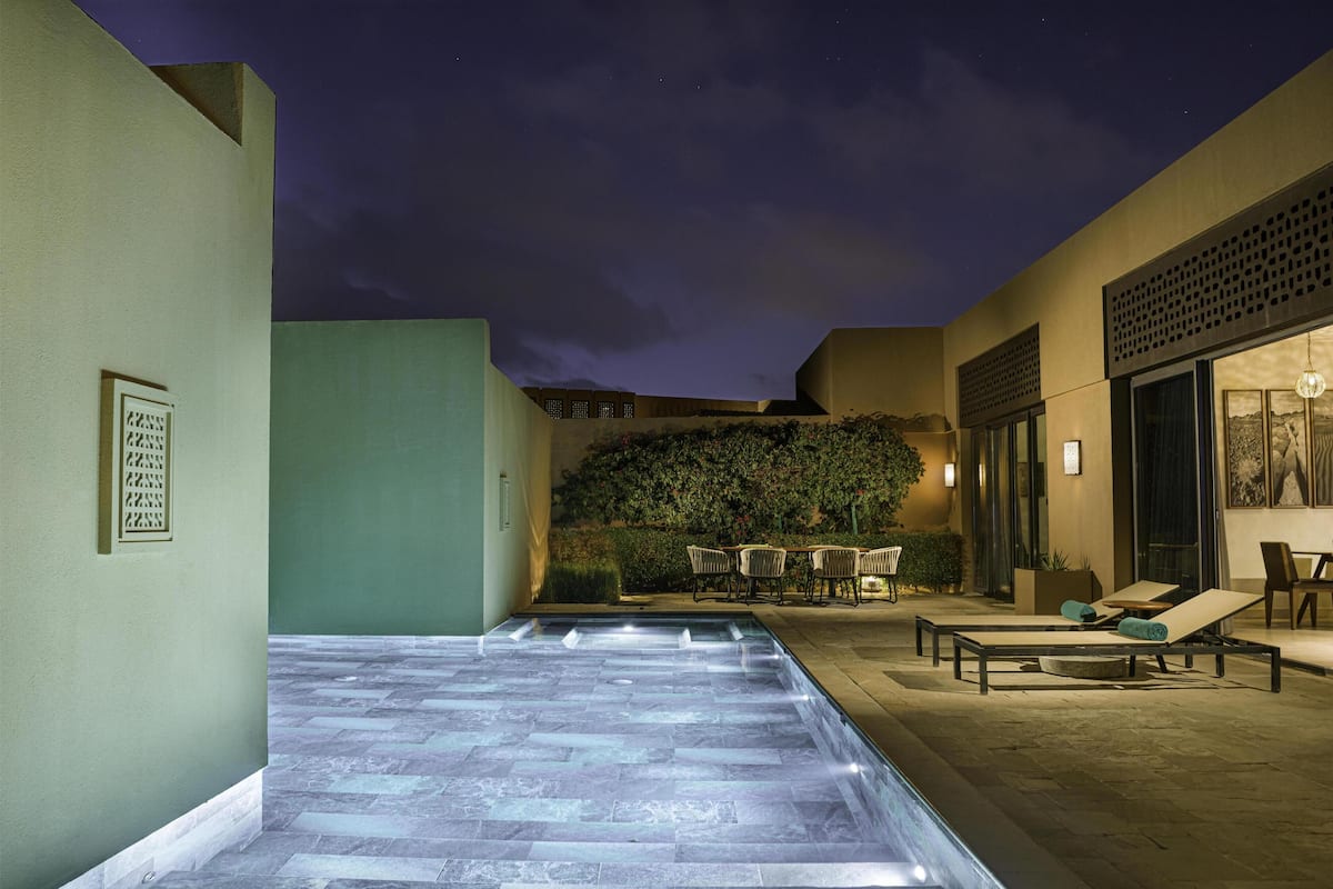 A nighttime outdoor pool area with a stone floor, lounge chairs, and a table with chairs.