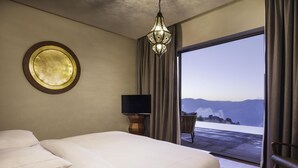Una camera d'albergo con un letto grande, una televisione su un mobile in legno, uno specchio rotondo appeso al muro e vista sulle montagne attraverso una porta scorrevole.