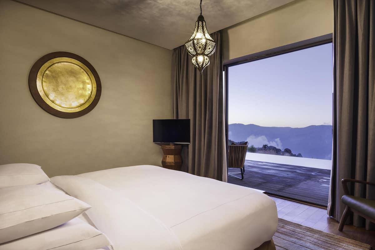 A hotel room with a large bed, a television on a wooden stand, a circular mirror on the wall, and a view of mountains through a sliding door.