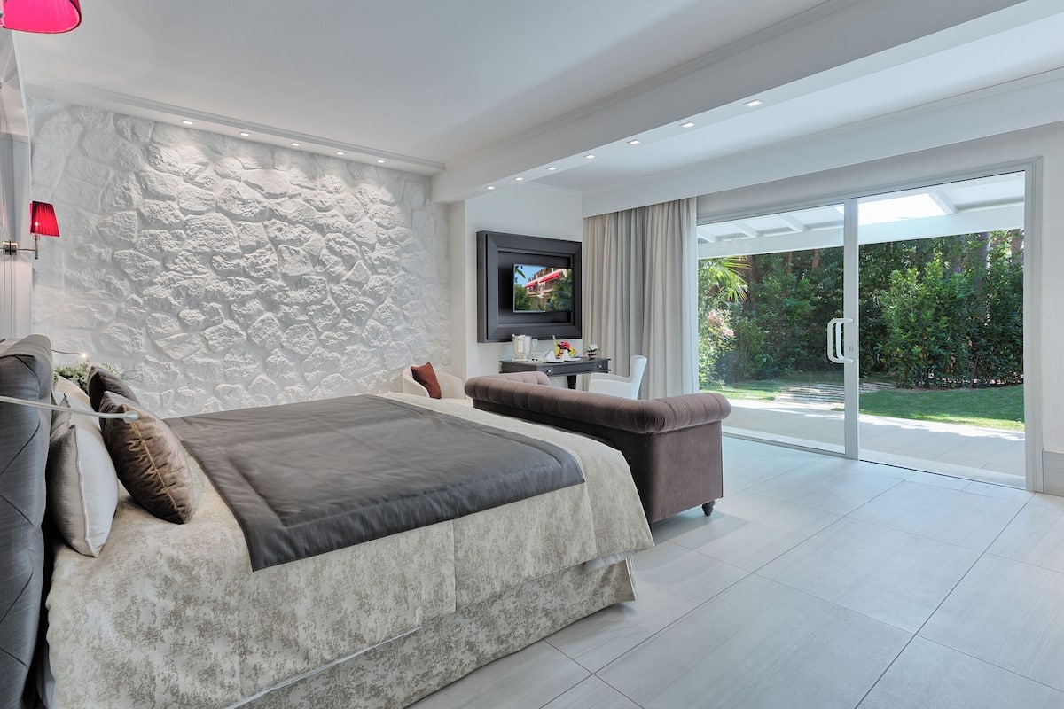 A modern bedroom with a stone wall, a bed, a desk, and a view of a garden through large sliding doors.