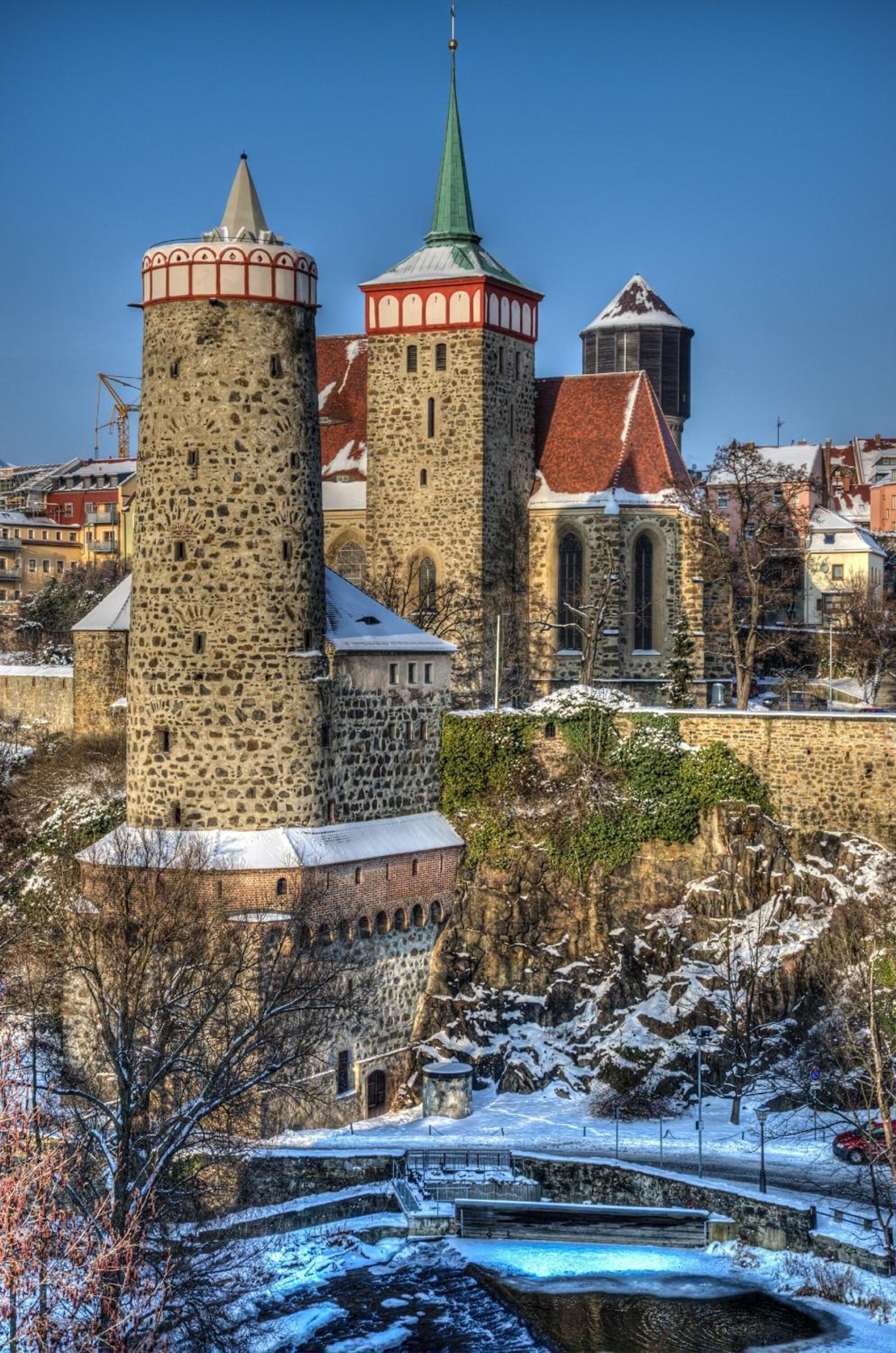 A historic stone tower with a red roof, adjacent to a church with a green spire, surrounded by snow-covered terrain and a small water body.