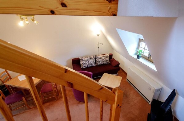 A loft-style room with a wooden staircase, a red sofa with patterned cushions, a small table, and a window with a plant. - Zum Handtuch (Bautzen)