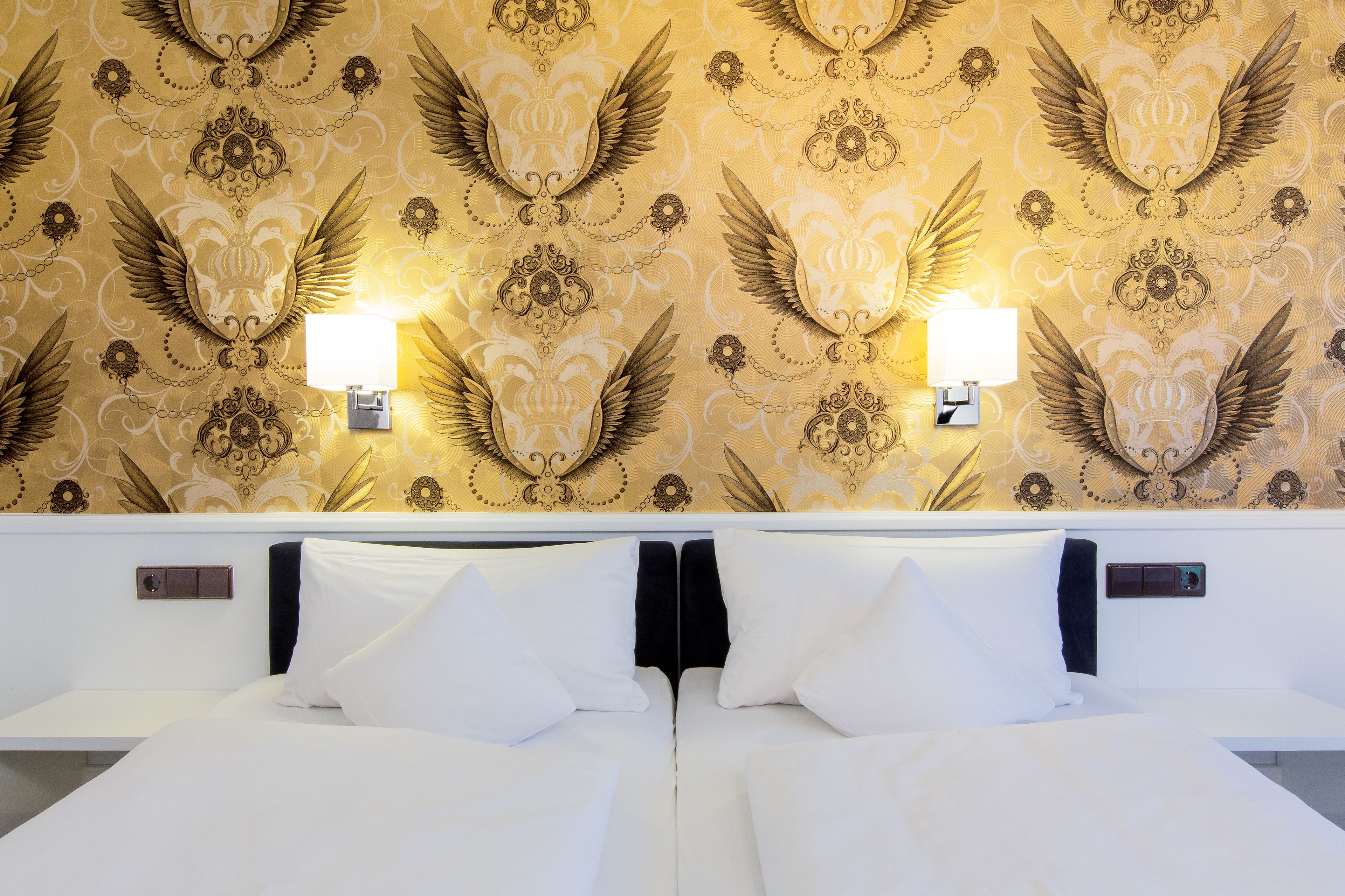 A hotel room with a large bed, two wall-mounted lamps, and a patterned wallpaper with wing motifs.