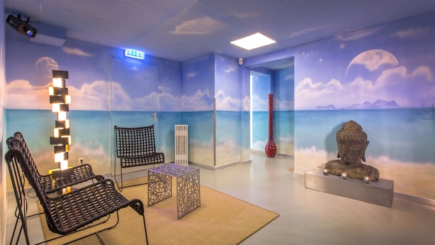 A room with a beach-themed mural, a black woven chair, a small table, and a statue on a pedestal. - Hotel Hellers Twenty Four 2 (Friedrichshafen)