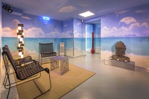Une pièce décorée d’une fresque murale sur le thème de la plage, avec une chaise en osier noire, une petite table et une statue posée sur un piédestal.