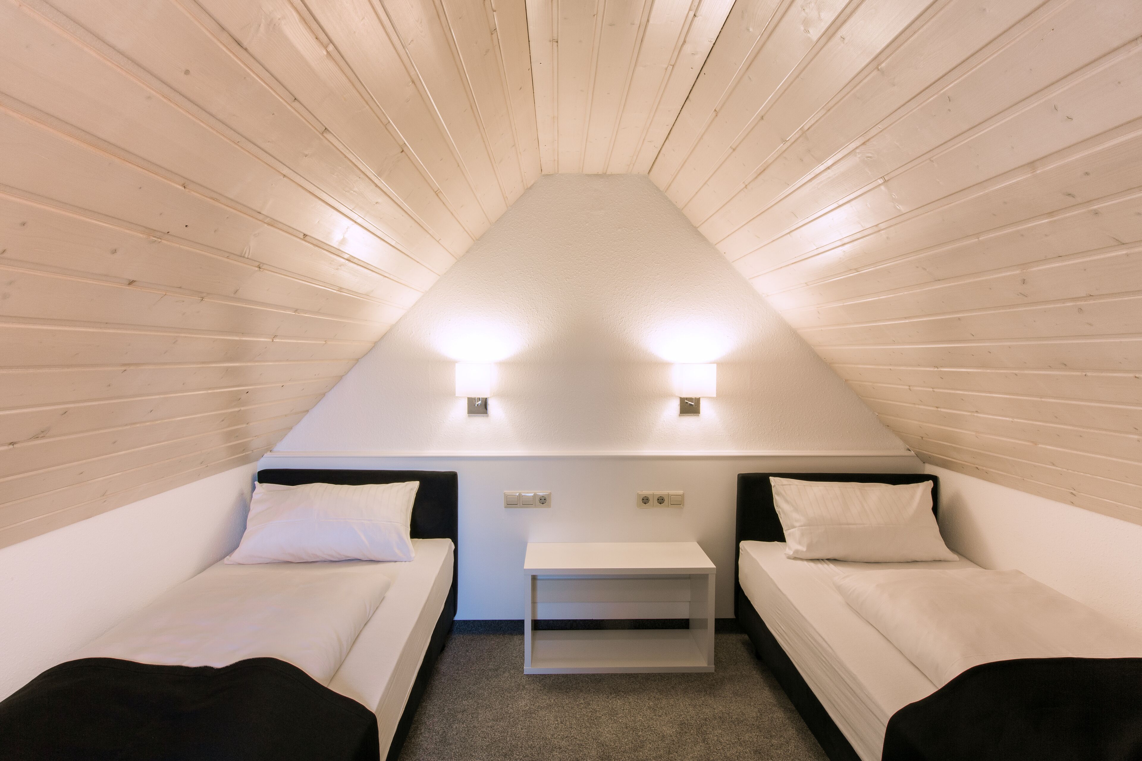 A small, triangular room with two single beds, a white nightstand, and two wall-mounted lights.