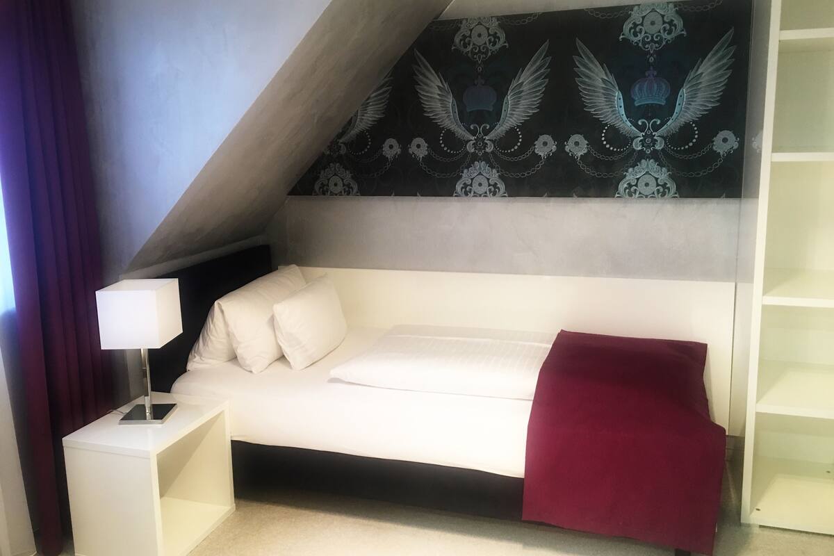 Un dormitorio con una cama blanca, una manta roja, una mesita de noche con lámpara y una pared con diseño decorativo.