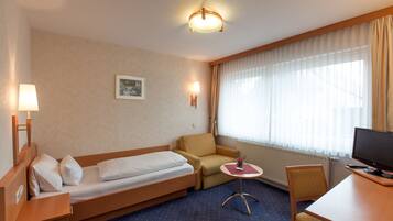 Ein Hotelzimmer mit einem Einzelbett, einem Sessel, einem kleinen Tisch, einem Schreibtisch mit Computer und einem Fenster mit Vorhängen.