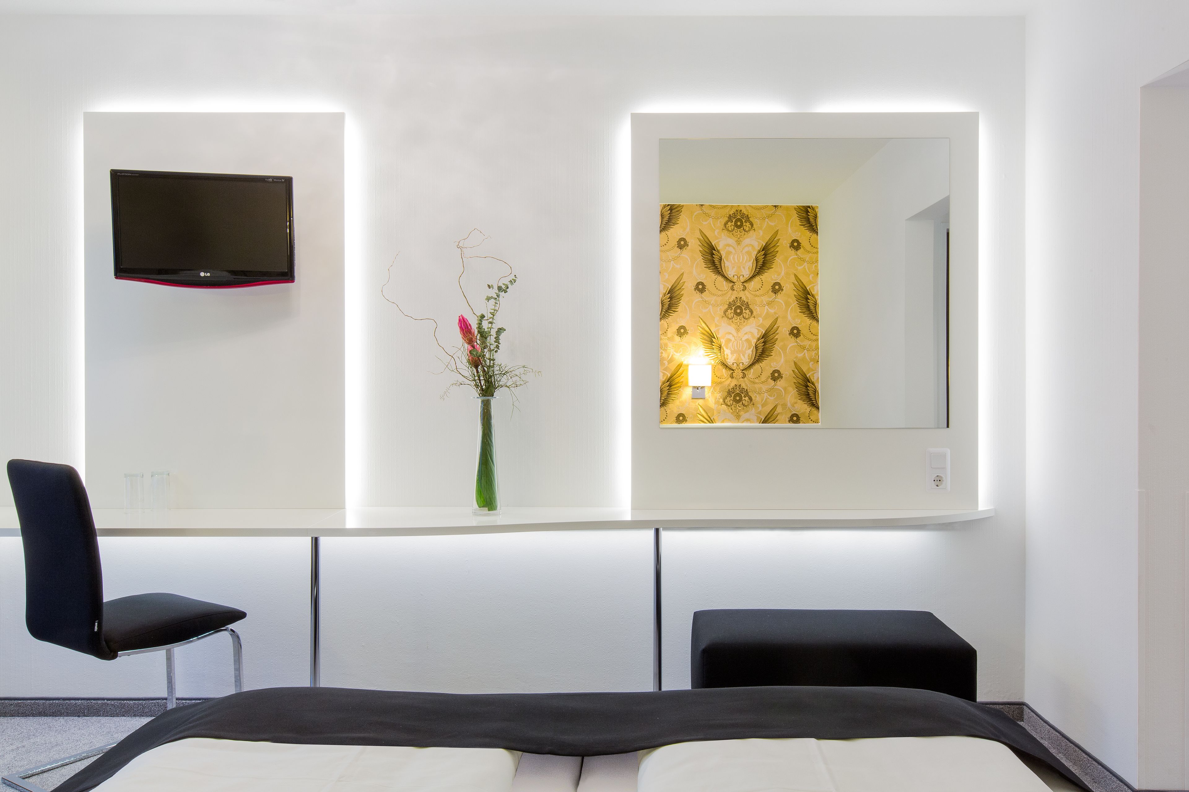 A modern hotel room with a flat-screen TV mounted on the wall, a bedside table with a vase of flowers, and a large mirror reflecting the room's decor.