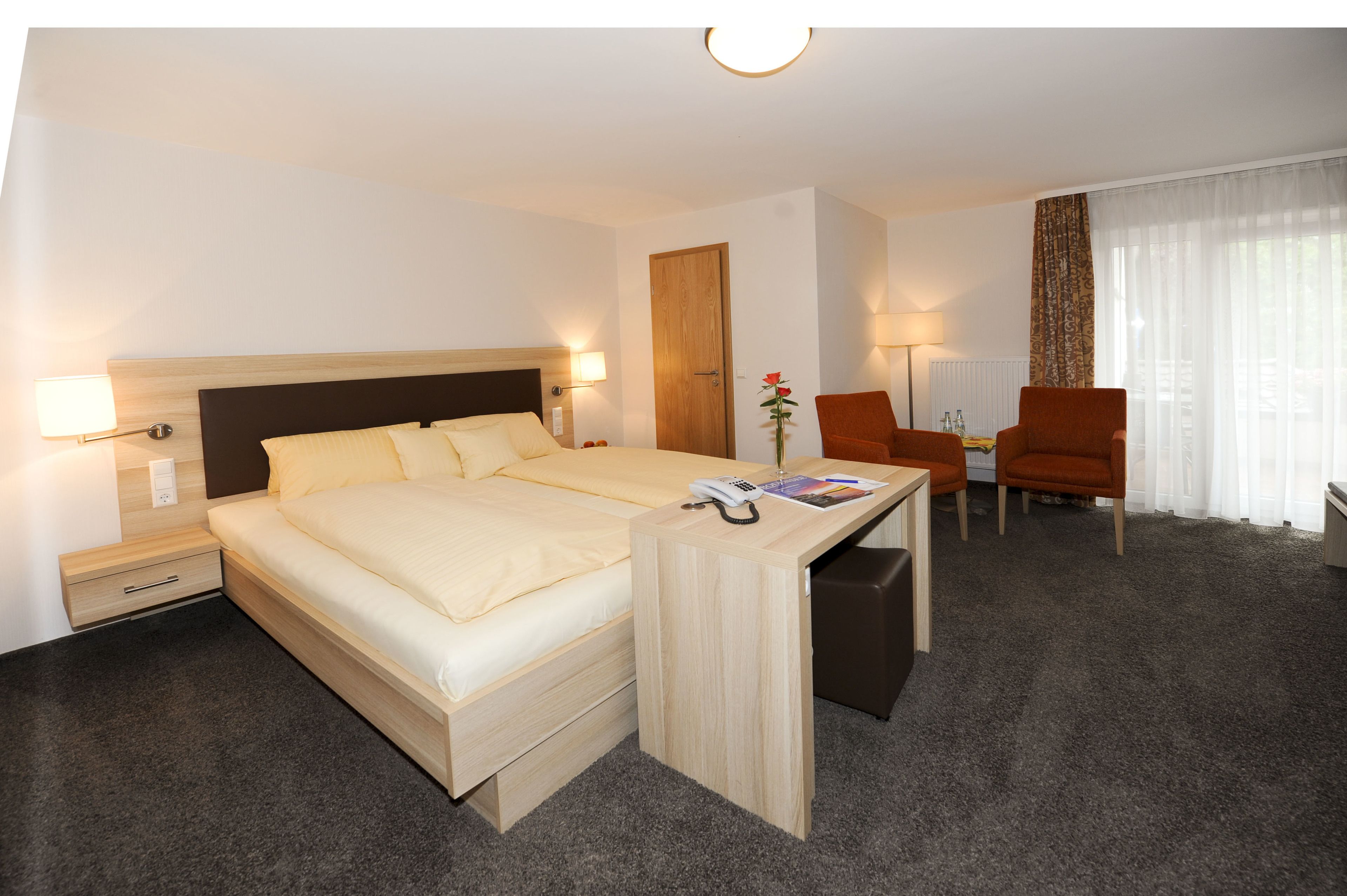 A hotel room with a bed, bedside table, lamp, chair, and a desk with a telephone and magazine.