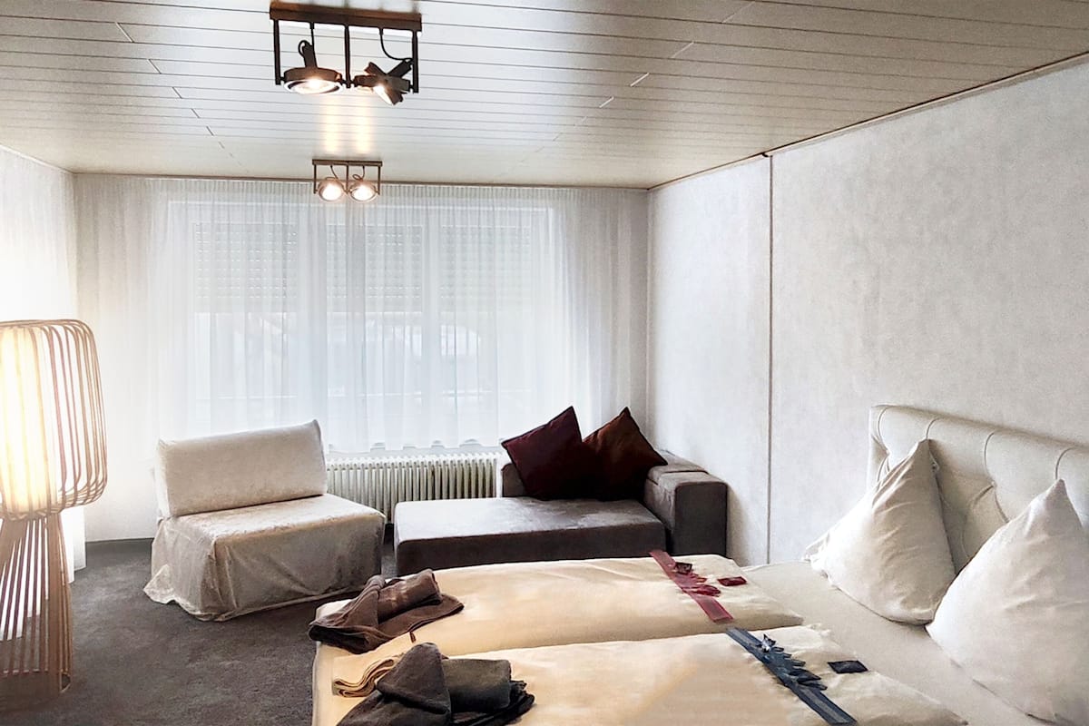 Habitación de hotel con cama, dos sillas, lámpara y ventana con cortinas traslúcidas.