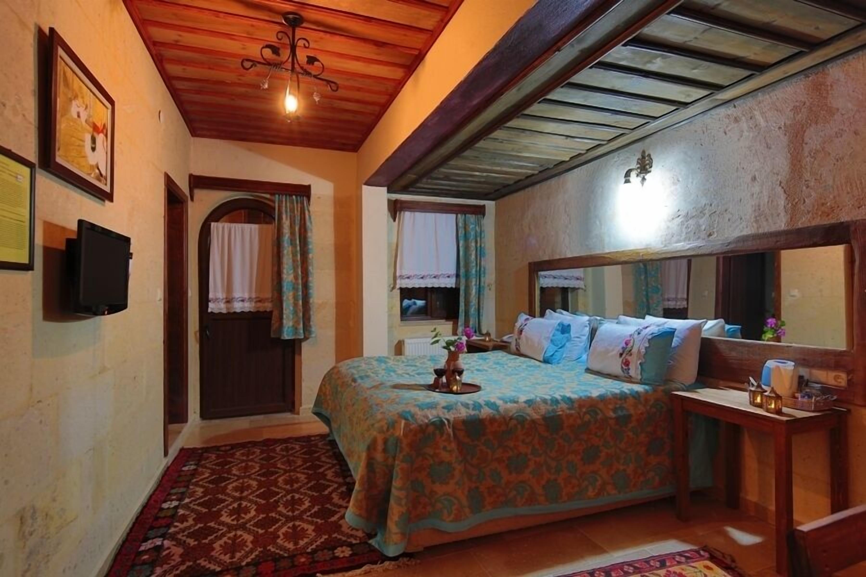 Un dormitorio con techo de madera, cama con colcha floral, mesita de noche, espejo y puerta con cortina.
