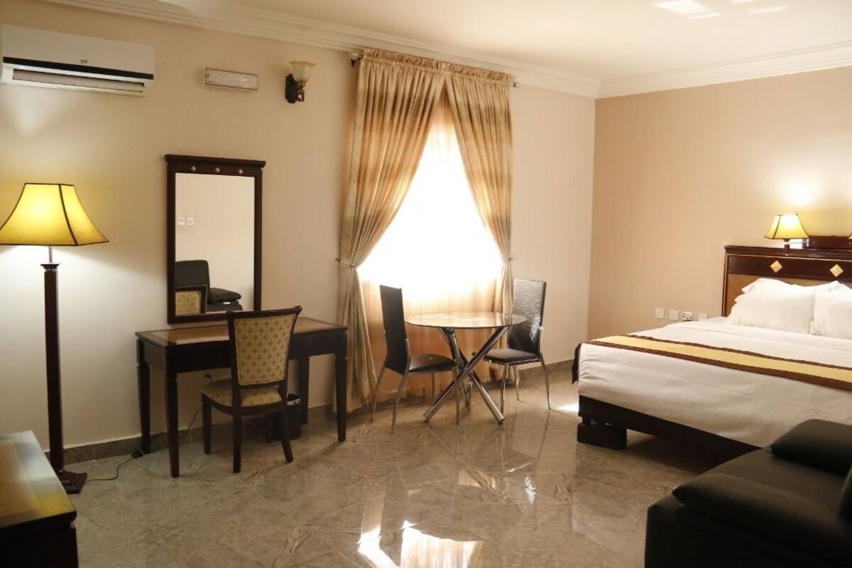 A hotel room with a bed, a desk, a chair, a table, a lamp, and a mirror.