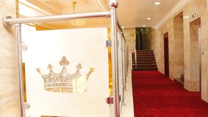 A hallway with a red carpet, a metal railing with a crown logo, and a staircase in the background. - Barca-Liga Hotels (Abuja)