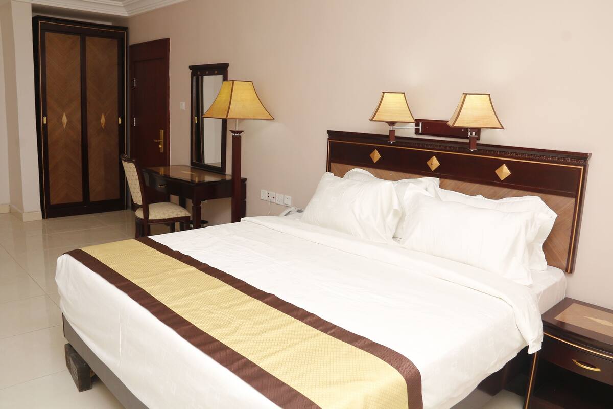 A hotel room with a large bed, bedside lamps, a desk with a chair, and a wardrobe.