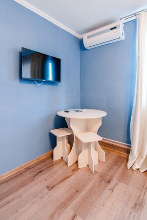 Quarto com parede azul, televisão de tela plana na parede, mesa redonda branca com dois banquinhos e unidade de ar condicionado sobre a mesa.
