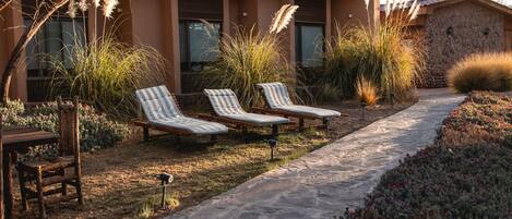 A modern building with a patio area featuring two lounge chairs, a table, and a pathway lined with plants.
