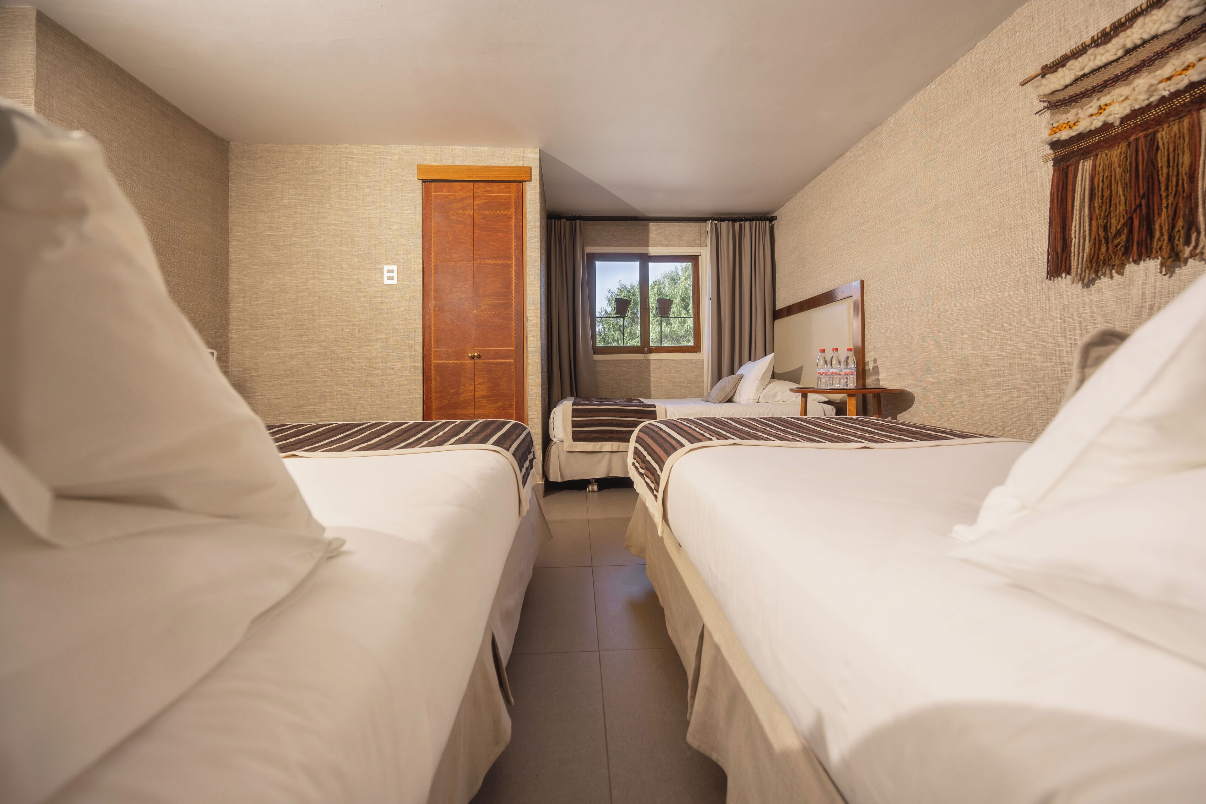 Ein Hotelzimmer mit zwei Betten, einer Holztür, einem Fenster mit Vorhängen und einem kleinen Tisch mit Flaschen.