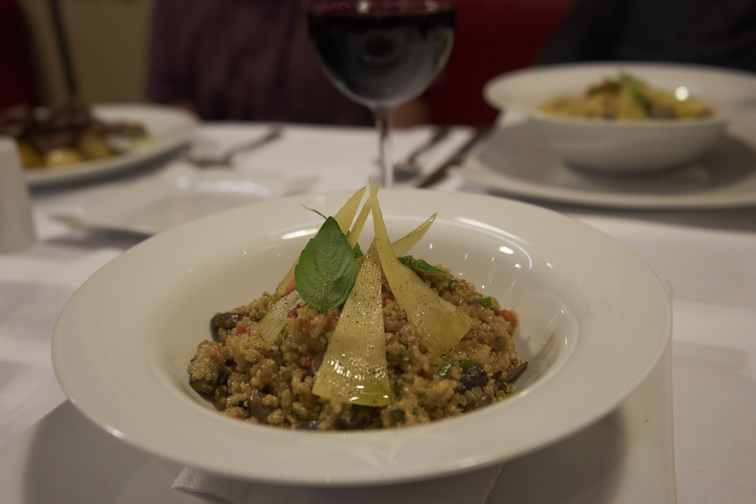 A white plate with a dish of food, a glass of red wine, and another plate in the background.