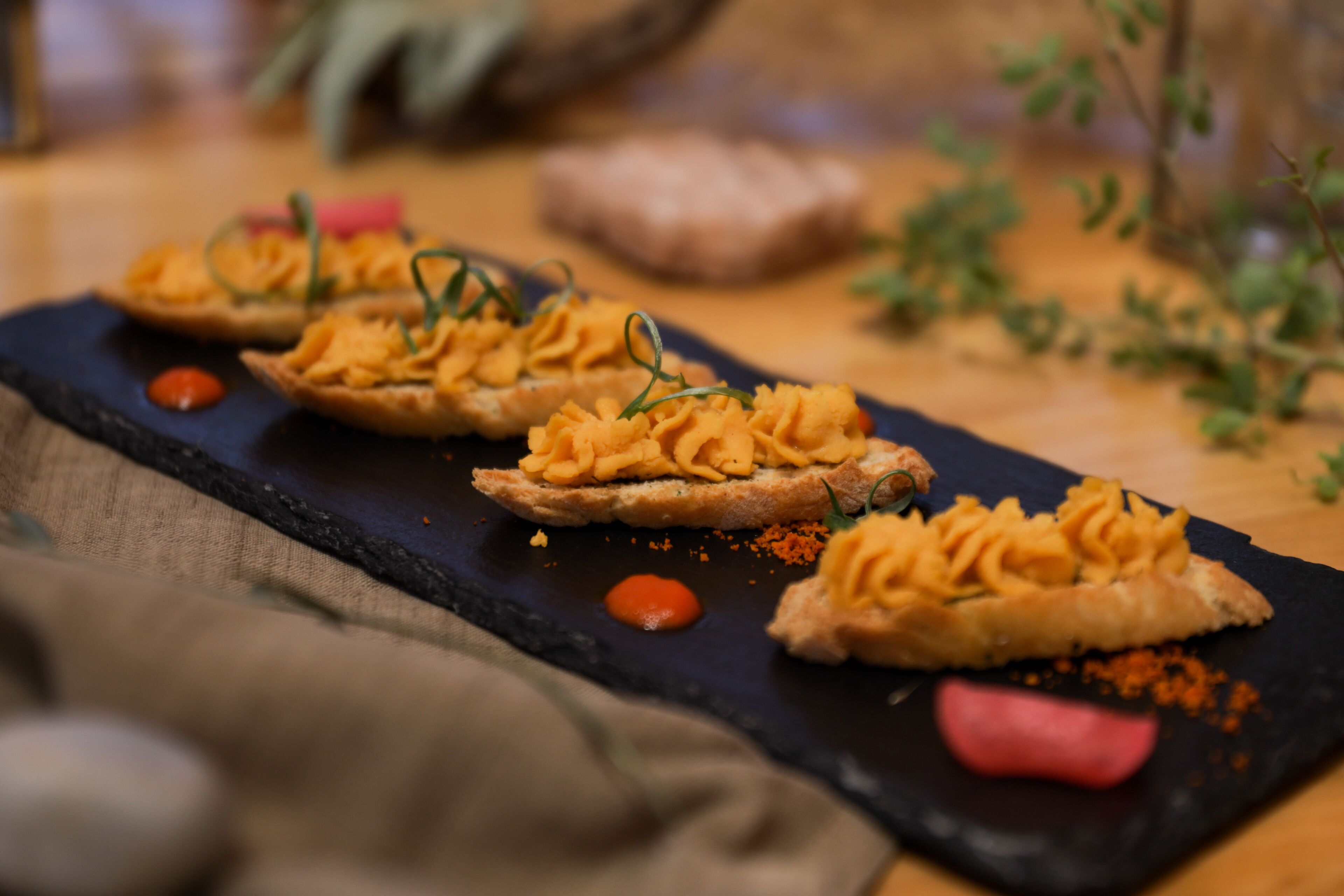 Une planche de service en ardoise noire, sur laquelle sont disposés trois morceaux de pain recouverts d’une pâte crémeuse, décorés d’herbes vertes et accompagnés d’une petite cuillerée de sauce rouge.