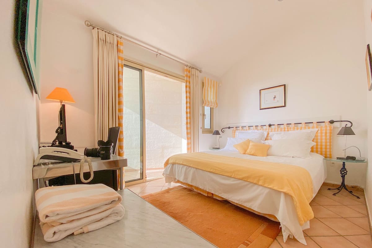 Ein Hotelzimmer mit einem Bett, einem Schreibtisch mit Telefon, einem Sessel, einem Fenster mit Vorhängen, einem Nachttisch mit Lampen und einem gerahmten Bild an der Wand.