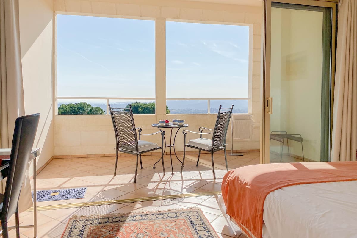 Ein Balkon mit zwei Stühlen und einem Tisch, ein Bett mit orangefarbener Bettdecke und Blick auf Grünflächen und ferne Hügel.
