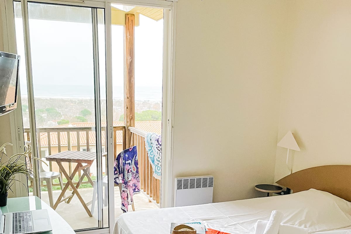 Une chambre d’hôtel avec un lit, un bureau avec un ordinateur portable, un chapeau posé sur le lit et un balcon avec vue.