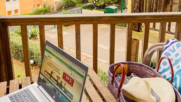 Een laptop op een houten tafel, met zicht op een kustplaatsje en een boom op de achtergrond.