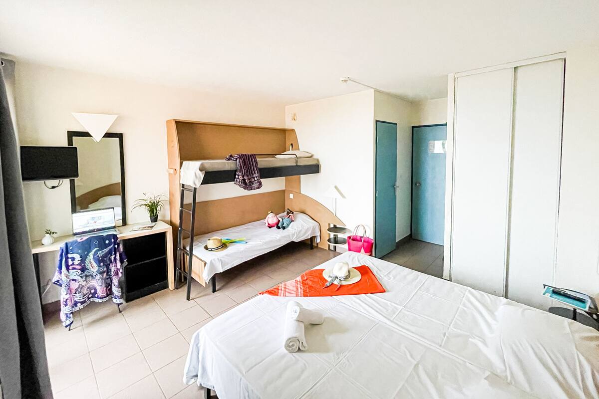 Une chambre d’hôtel avec un lit superposé, un bureau avec un ordinateur, un miroir, une table de chevet avec une lampe et une armoire.