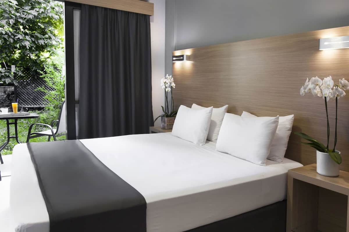 A hotel room with a large bed, a wooden headboard, a small table with a chair, and a potted plant.