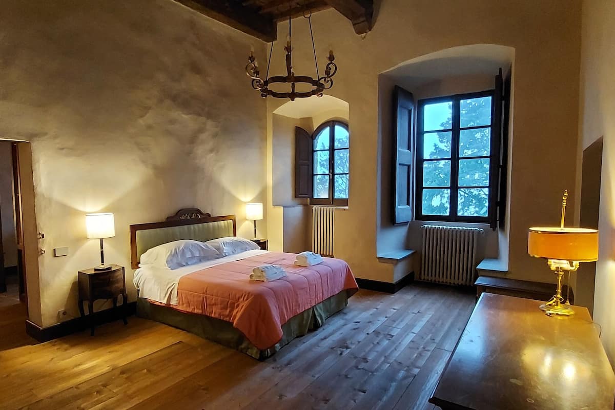 Un dormitorio con piso de madera, una cama con cubrecama rosa, dos lámparas de noche, un candelabro, una ventana con contraventanas y un radiador.