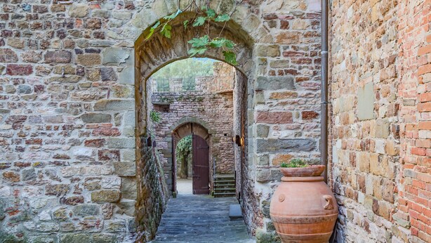 A narrow stone corridor with an arched entrance leading to a wooden door, a large terracotta pot, and a brick wall. - Castello di Mugnana (Greve in Chianti)