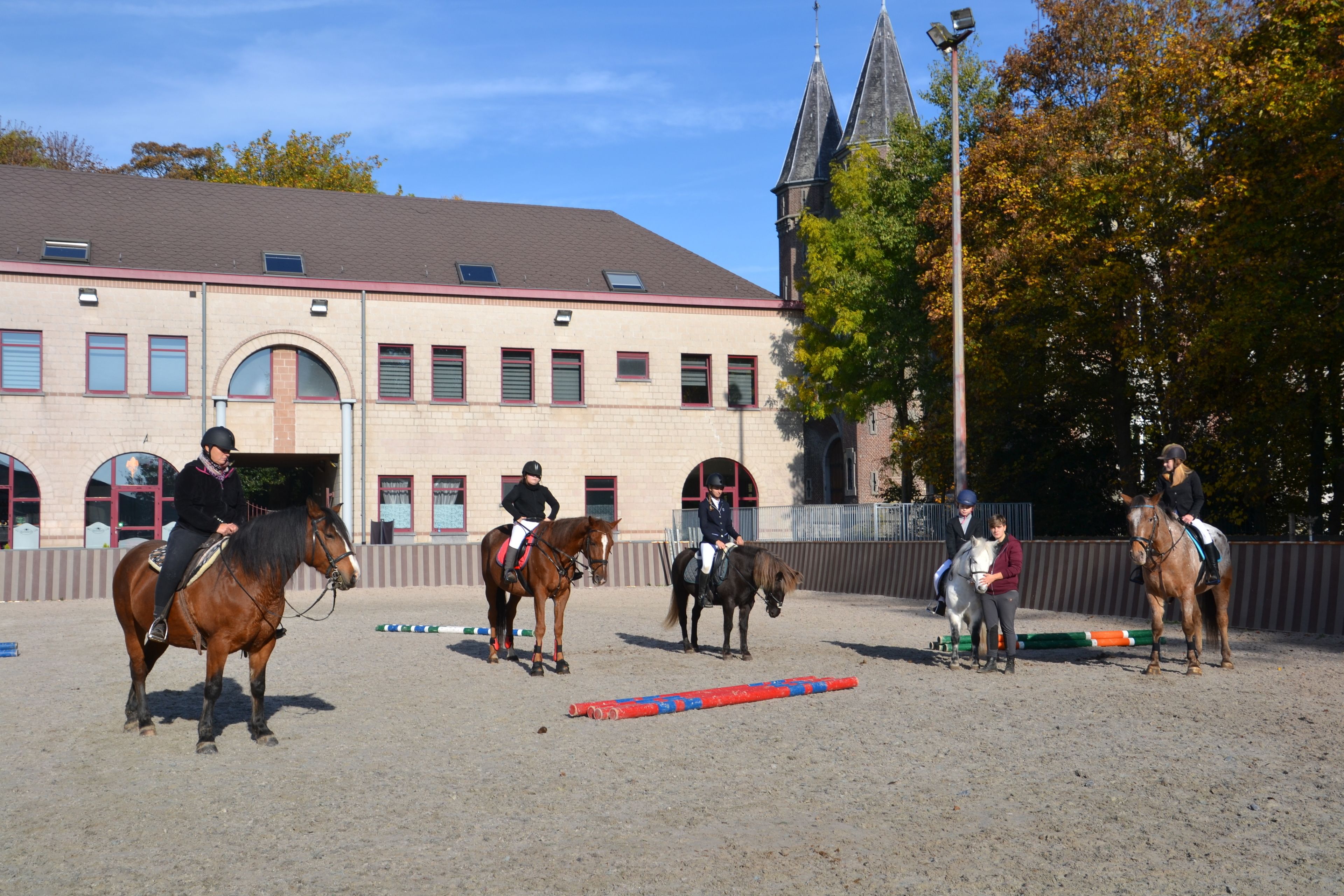 An indoor riding arena with horses and riders, a building with arched windows, and a church steeple in the background.