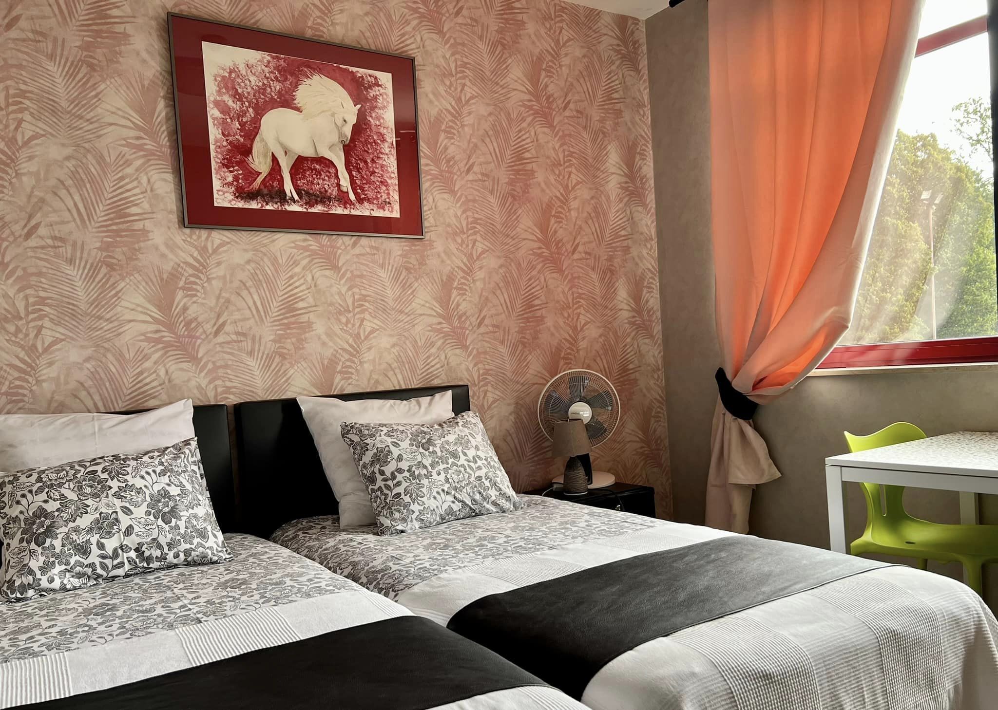 Une chambre avec deux lits, un tableau encadré représentant un cheval, un ventilateur sur pied et une petite table avec une chaise.