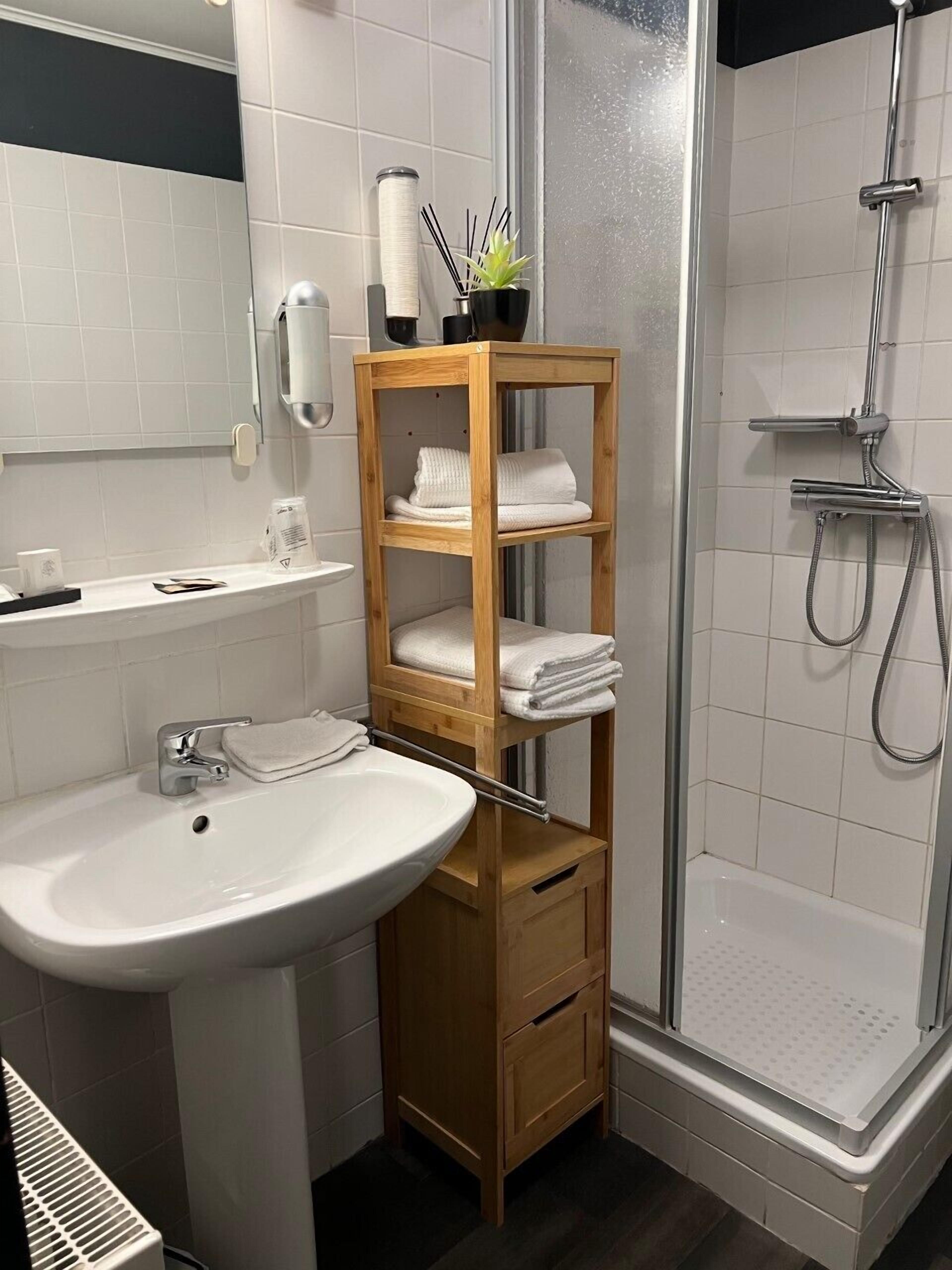 Une salle de bain avec un lavabo blanc, une étagère en bois avec des serviettes, une douche et des toilettes.