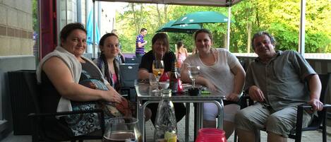 Un grupo de personas sentadas en una mesa al aire libre, con bebidas y una vela, bajo una estructura cubierta y con vistas a árboles.