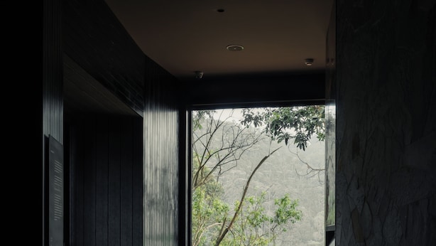 A modern interior with a dark wood floor, a seating area with cushions, a round table, and a large window with a view of greenery. - Silverback Lodge (Bwindi)