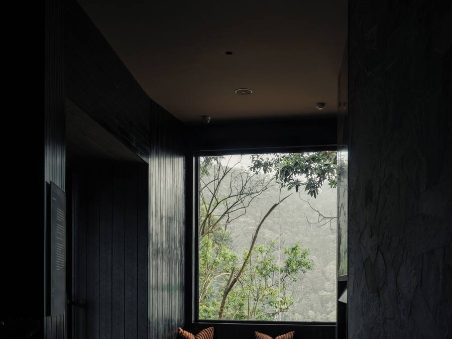 A modern interior with a dark wood floor, a seating area with cushions, a round table, and a large window with a view of greenery.