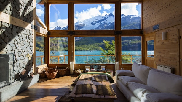 A cabin with large windows offering mountain views, a stone wall, and a cozy seating area with a fireplace. - Aguas Arriba Lodge (El Chalten)