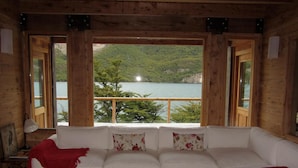 A cozy cabin with a white sofa, a coffee table with books, and a scenic view of a lake and mountains through large windows. - Aguas Arriba Lodge (El Chalten)