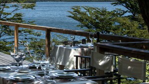 A dining table set with plates, glasses, and cutlery, overlooking a lake with mountains in the background. - Aguas Arriba Lodge (El Chalten)