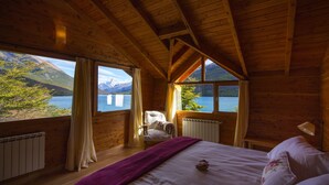 A wooden cabin bedroom with a large bed, a chair, and a view of a lake and mountains. - Aguas Arriba Lodge (El Chalten)