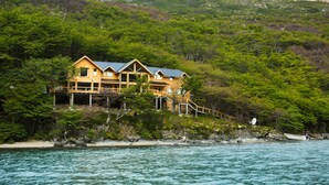 A wooden house with a balcony, surrounded by lush greenery and a body of water. - Aguas Arriba Lodge (El Chalten)