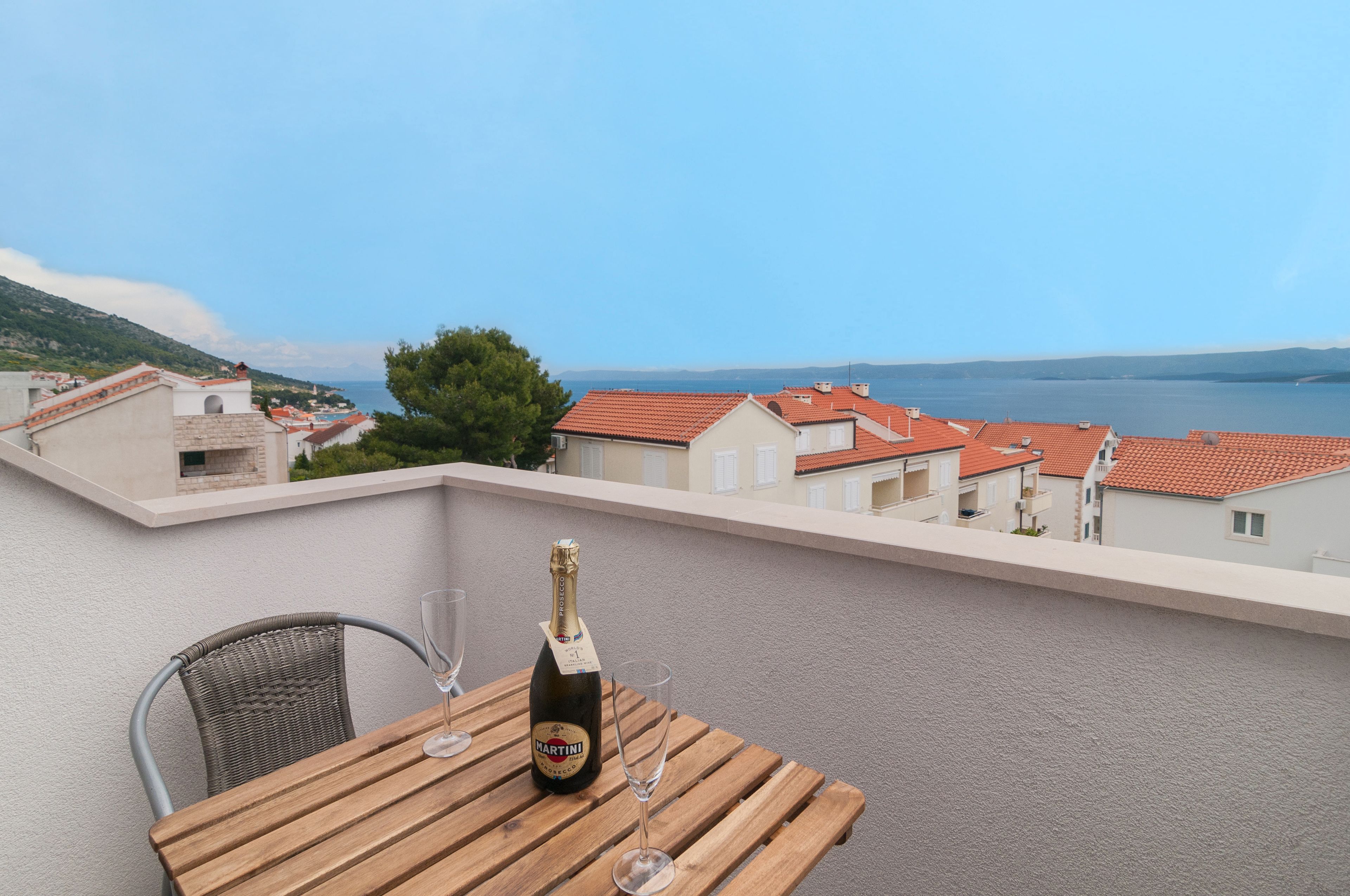 A balcony with a wooden table, a bottle of Martini champagne, and two glasses. The view includes red-roofed buildings and a body of water.
